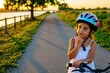 © Viktor - Little girl wears colorful cycling helmet riding bike on sunny park path. Thoughtful preschool child touches chin looking away during outdoor activity. Cute kid stands with bicycle in nature at