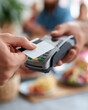© Tina - Person taps card on payment terminal. Focus on hands and device, blurred background. Dish and drink suggest casual dining. Scene implies modern, everyday commerce. People's emotions unclear