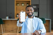 © Daniel - Smiling man seated in a modern kitchen holds a smartphone toward the camera with a blank white screen, perfect for app mockups, tech presentations, social media promotion, and remote