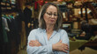 © Krakenimages.com - Hispanic senior woman with grey hair and glasses stands with arms crossed and necklace visible among clothing racks in building; frustration.
