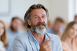 © lashkhidzetim - Bearded man speaks during professional seminar. Focused audience listens attentively behind him. Warm, engaging expression conveys confidence and passion. Ideal for business, education