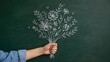© MSTLIMA - A hand holding a beautiful bouquet of wild flowers drawn in white chalk on a dark black chalkboard surface representing a creative artistic gift for a special occasion or holiday event