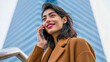 © Eva Corbella - Charming woman with radiant smile chatting on her smartphone against a striking modern building in bright daylight