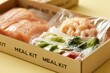 © Marina Demidiuk - Raw salmon fish fillet, shrimps and green vegetables inside vacuum plastic bags in cardboard box. Meal Kit Delivery, nutrition kits. Fresh food ingredients for cooking. Home wellness. Ready made set