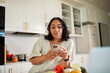 © Marko Geber - Young woman using smartphone while cooking in home kitchen