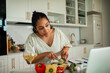 © Marko Geber - Young woman cutting cucumber while watching laptop in modern kitchen