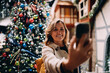 © BullRun - Portrait of smiling mature woman looking at smartphone screen to take selfie, happy female posing with mobile phone near Christmas tree, digital communication, authentic representation, tech