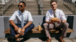 © pattozher - Lunch Break with Colleagues: Two businessmen, dressed in smart office attire, enjoy a casual lunch break outdoors, sitting on a bench with sandwiches and drinks.