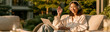 © pattozher - Outdoor Work Session: A young Asian woman wearing headphones is comfortably seated on a patio sofa, engaged in a video call on her laptop, bathed in the warm, natural light of the outdoors.