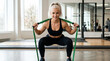 © Len_mik - Smiling blonde woman performing squats with a green resistance band in a bright gym. Happy female athlete doing lower body strength training. Fitness and active lifestyle concept