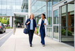 © Kassiopeia  - A woman in scrubs and a lab coat walks beside a woman with a bag. They are leaving a hospital. The sun shines in the afternoon