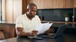 © LA Stock Vision - Black man reviewing financial paperwork and utility bills at home with a laptop.