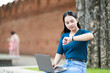 © Tj - Young Asian woman glancing at her fitness tracker while using a laptop to work remotely in a park, balancing a travel lifestyle with her digital nomad profession