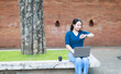 © Tj - Young Asian woman sitting outdoors with a laptop and coffee, checking her watch, embodying digital nomad lifestyle and time management for remote work and freelance