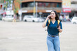 © Tj - Young asian woman walking along an urban street, talking on her smartphone while holding a takeaway coffee, wearing a backpack for travel or daily activity