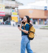 © Tj - Young Asian woman traveling, carrying a backpack and holding a takeaway coffee, engaging in a phone conversation while walking through a vibrant urban environment