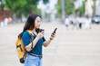 © Tj - Young Asian woman traveler carrying a backpack, sipping coffee from a disposable cup, and actively looking at her smartphone for navigation or communication while exploring a city