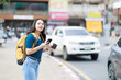 © Tj - Young Asian woman holding smartphone and coffee cup, wearing backpack, navigating on city street, looking for transportation or a ride-sharing pickup