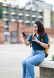 © Tj - Asian female traveler enjoying a city break, looking at her mobile phone, carrying a backpack, and drinking coffee while sitting on a stone bench in front of the Tha Phae Gate