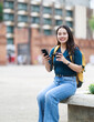 © Tj - Young Asian woman traveler sitting on a stone bench in a city square, looking at the camera, holding a smartphone and takeaway coffee cup, enjoying her solo adventure outdoors
