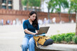 © Tj - Young female student sitting outdoors at Tha Phae Gate in Chiang Mai, Thailand, studying online and working on her laptop with a book and backpack nearby