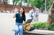 © Tj - Young Asian woman sitting outdoors, holding a laptop and backpack, waving hand, greeting or saying goodbye, enjoying urban university life and travel abroad