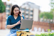 © Tj - Young Asian woman student studying online, talking on mobile phone and using laptop for distance learning or remote working in an urban city park
