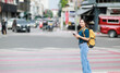 © Tj - Backpacker woman standing on city street crossing, smiling and using mobile phone, navigating urban adventure while exploring a Thailand destination