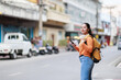 © Tj - Young asian woman traveler carrying a backpack and headphones. Holding smartphone. Looking at the camera. Exploring a vibrant Southeast Asian city street