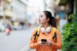 © Tj - Young Asian woman exploring an urban street, wearing headphones around her neck and carrying a backpack, using a mobile phone while looking forward