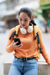 © Tj - Young adult woman holding a smartphone and wearing a backpack with headphones around her neck, engrossed in digital connectivity and communication outdoors