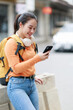 © Tj - Young smiling Asian woman wearing orange long sleeve shirt, light blue jeans, and a yellow backpack, sitting on a concrete barrier and using her smartphone on a city street