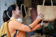 © Tj - Young woman shopping for handmade items in a traditional store, happily choosing a straw bag, reflecting concepts of travel, local crafts, and tourism