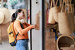 © Tj - Young Asian woman traveler with backpack and headphones shopping for handmade straw bags at a street market, enjoying her leisure time and vacation lifestyle