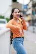 © Tj - Young Asian woman smiling, standing outdoors, holding and talking on a mobile phone, wearing an orange long-sleeved shirt, blue jeans, and a backpack, communicating in an urban setting