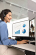 © fizkes - Businesswoman with headset sits at office desk, participating in video call, reviewing project analytics on dual-monitor, interacts with diverse colleagues, discussing charts, analyzes data on screen