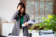 © Matawut - Asian businesswoman feeling overwhelmed and stressed from work, suffering a headache while carrying a stack of documents in a busy office environment, experiencing burnout