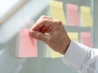 © zabhie - Businessman Placing Sticky Note on Glass Wall During Brainstorming Session, Close Up of Professional Hand Organizing Project Task List with Colorful Notes in Office