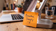 © alex - An orange sticky note reading 'Breathe Deeply' is attached to a metallic desk lamp base next to an open laptop and a glass of water on a wooden desk.