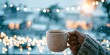 © Bonsales - Hand holding warm ceramic mug outdoors during gentle snowfall, celebrating winter holiday spirit with glowing lights