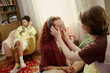 © DragonImages - Young Caucasian woman applying eye patches on young girl during home skincare routine, while Black teenager using smartphone nearby. Ideal for beauty self care and friendship marketing