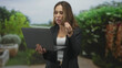 © Krakenimages.com - Woman holding laptop and gesturing with right hand during a video call in a garden patio with potted plants and walkway; focus remote meeting.