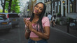 © Krakenimages.com - Black woman clapping hands on a street wearing pink crop top and jeans, braided hair and bracelets while smiling at camera; confidence warmth.