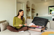 © Home-stock - Pregnant woman sitting on sofa beside suitcase filled with baby clothes, writing checklist in notebook, preparing for hospital stay