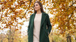 © Emre Akkoyun - Woman enjoying autumn park, wearing green coat and sweater, looking away from camera with fall foliage background