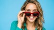 © Ali - Glamorous young woman in teal blouse peeking playfully over bold red sunglasses, smiling brightly, cheerful summer fashion portrait against vibrant blue background, lifestyle beauty shot