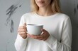 © Maryna - Young woman holds white mug with hot drink. Female enjoys beverage, maybe coffee or tea, in cozy white sweater. Casual morning indoors moment.