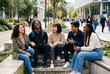 © Kassiopeia  - Five friends sit on steps in a city area and laugh together. They enjoy their time and share stories. People walk by in the background. The sun shines brightly