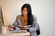 © Prathankarnpap - Asian businesswoman reading notebook at office desk with computer monitor and coffee mug, focused and professional