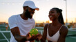© dinatychynska - A smiling couple exchanges a pickleball before the next point, warm sunset light and string lights glowing on the court behind them. A romantic summer lifestyle scene of connection and play.